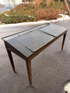 Antike Schulbank / Schultisch ca. 1900-1930 - mit Patina. Originale antike Schulbank aus Holz, vermutlich aus der Zeit ca. 1900-1930. Klassischer Schultisch mit zwei Klappdeckeln und integrierten Ablagefächern, wie sie früher in Schulzimmern verwendet wurden.
Die Tintenloch-Öffnungen sind noch sichtbar.
Der Tisch weist eine wunderschöne, authentische Patina mit altersbedingten Gebrauchsspuren auf. Die schwarze Lackierung ist teilweise stark gekrakelt, was den historischen Charakter unterstreicht.
Zustand:
• Beide originalen Klappdeckel sind vorhanden
• Die Scharniere sind ausgerissen, können jedoch fachgerecht repariert werden
• Holzsubstanz insgesamt stabil
• Alters- und Gebrauchsspuren gemäss Fotos
Ideal als Dekorationsobjekt, für ein Vintage-Interieur, Atelier, Ladenbau oder als Restaurationsprojekt für
Liebhaber alter Schulmöbel.
Masse: ca. 89 x 67 x 160